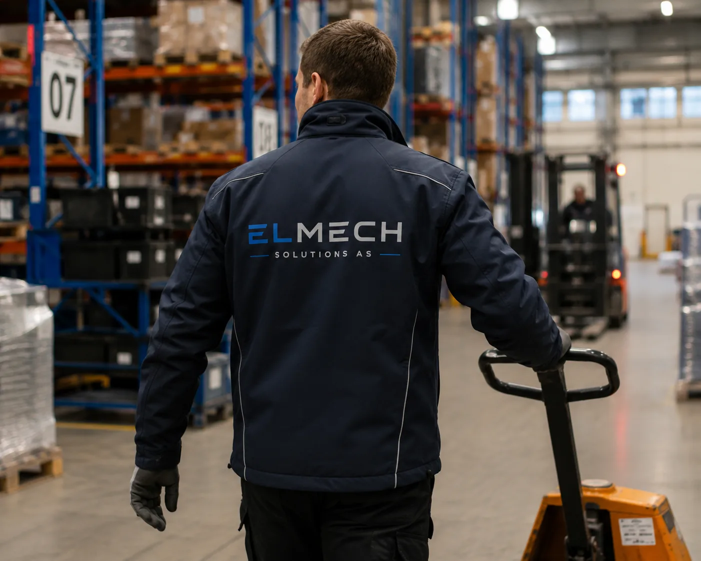 Tilsett sett bakfrå i ELMECH-jakke, dreg ein orange palletruck mellom høge lagerreoler