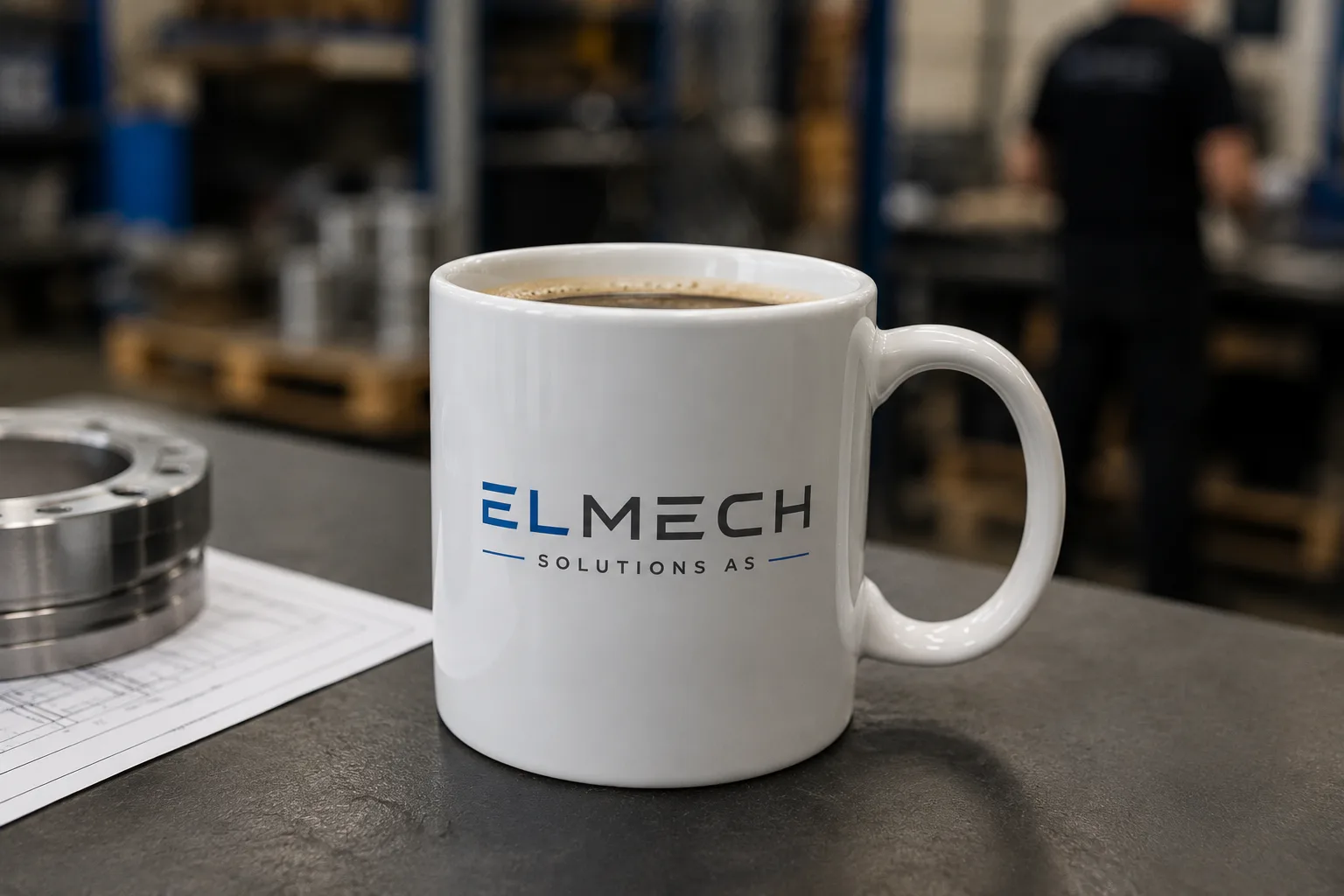 Kvit kaffekopp med ELMECH SOLUTIONS AS-logo, ståande på arbeidsbenk i industrimiljø